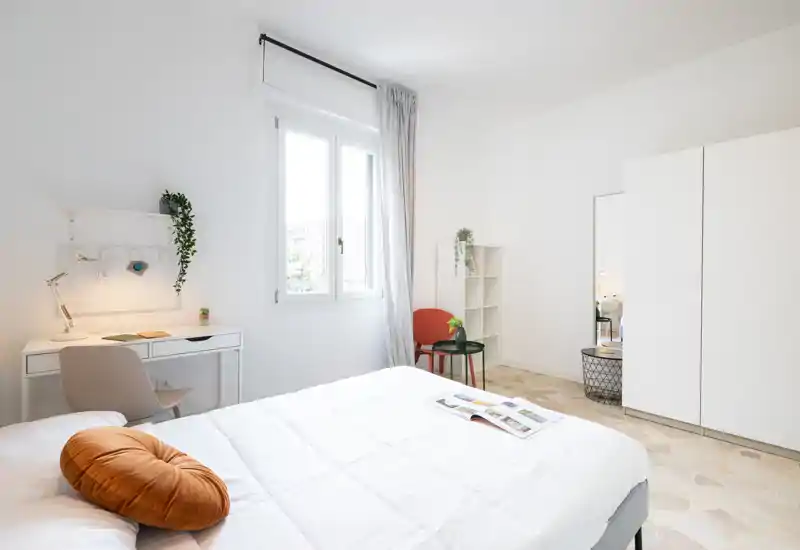 Dormitorio luminoso y minimalista con una cama blanca grande, textiles suaves y un pequeño escritorio junto a la ventana. La estancia es luminosa con tonos neutros, un cojín decorativo y plantas que aportan calidez.