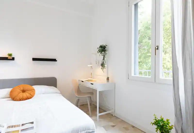Dormitorio luminoso y minimalista con una cómoda cama doble, un pequeño escritorio blanco junto a la ventana y plantas verdes que aportan frescura.