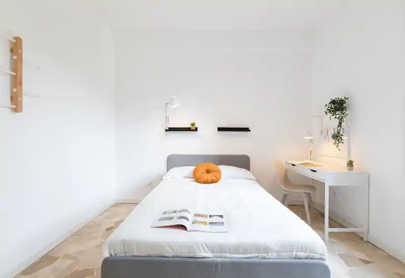 Dormitorio luminoso y minimalista con cama doble centrada, ropa de cama blanca y un cojín redondo cálido; a la derecha hay un pequeño escritorio con lámpara y planta que crean un rincón funcional y tranquilo.