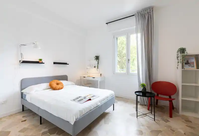 Dormitorio moderno y luminoso con cama doble bien hecha, lámpara auxiliar, pequeño escritorio junto a la ventana y detalles decorativos elegantes.