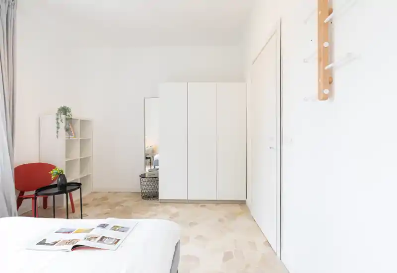 Dormitorio luminoso y minimalista con la cama visible en primer plano, armario blanco, estantería y una silla roja que crean un ambiente sencillo y acogedor.
