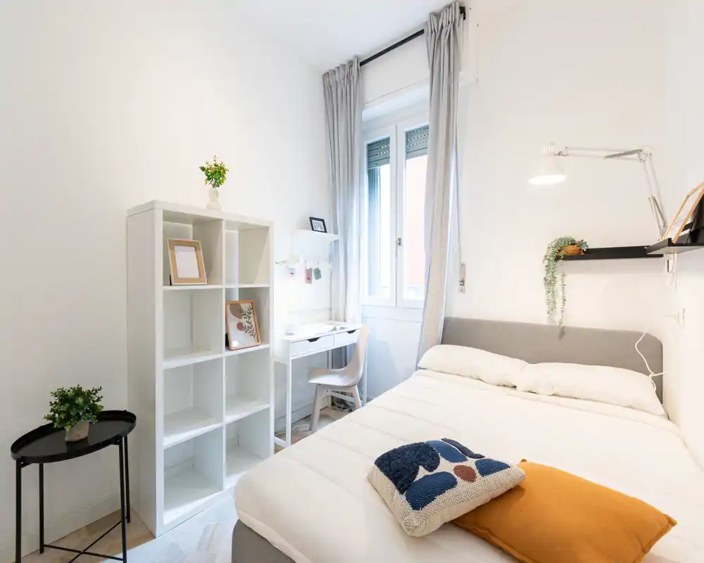 Camera da letto singola luminosa e minimal con letto comodo, scrivania vicino alla finestra e mensole eleganti — ideale per uno studente o professionista.