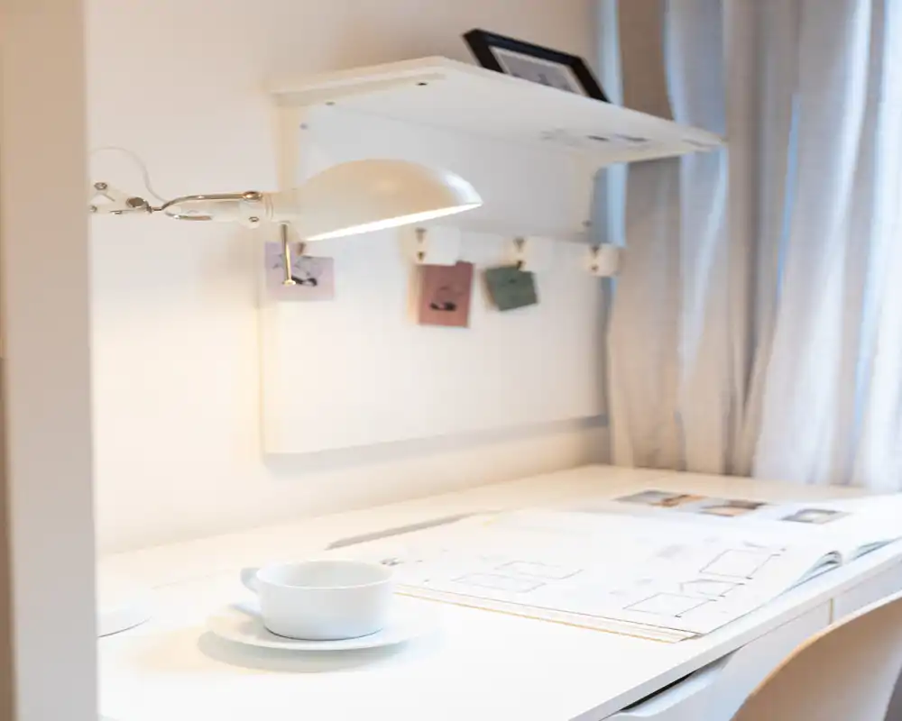 Angolo studio luminoso e minimal con scrivania bianca, lampada regolabile e una tazza su piattino — perfetto per leggere o lavorare in tranquillità.