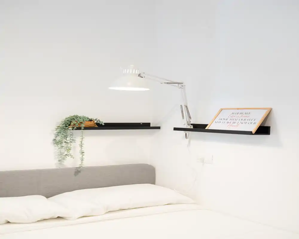 Camera da letto luminosa e minimal con letto ben fatto, mensole a parete e una lampada snodata che creano un’atmosfera moderna e tranquilla.