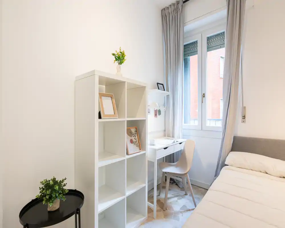 Camera da letto luminosa e minimal con letto singolo, scrivania compatta vicino alla finestra e libreria a cubi. La luce naturale e i toni neutri creano un'atmosfera moderna e rilassante, perfetta per un monolocale.