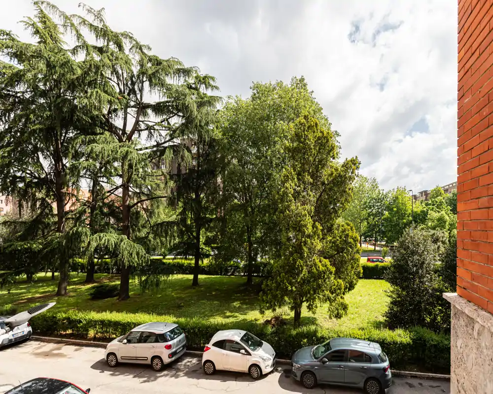 Vista a livello strada su un giardino condominiale verde con alberi maturi e auto parcheggiate in primo piano — ideale per mostrare lo spazio esterno e il verde del quartiere.
