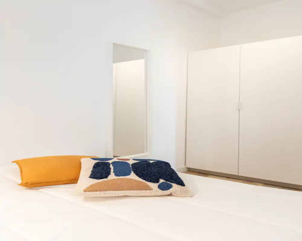 Camera da letto minimalista e luminosa con letto ordinato, cuscini decorativi testurizzati e un cuscino accentato color senape; pareti neutre e un ampio armadio creano un’atmosfera calma e ariosa.