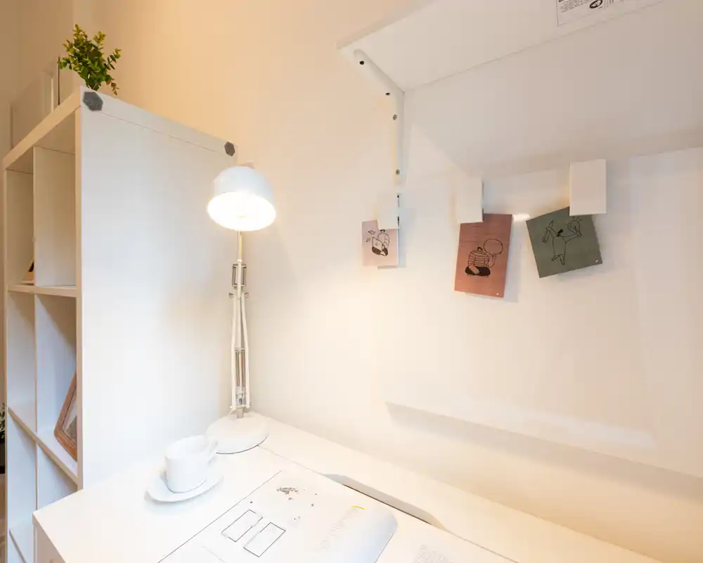 Angolo luminoso e minimal del soggiorno con scrivania bianca, lampada da tavolo, appunti decorativi e scaffalatura — uno spazio di lavoro pulito e moderno integrato nell'area giorno.