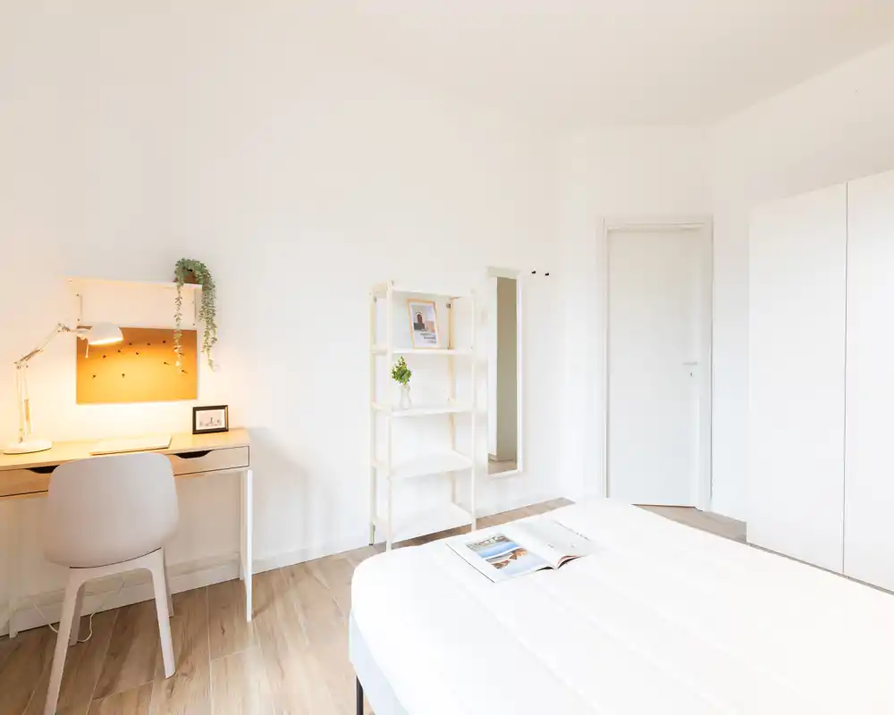 Dormitorio luminoso y minimalista con cama blanca en primer plano, escritorio con lámpara encendida, estantería y armario — aireado, moderno y bien presentado para anuncios.
