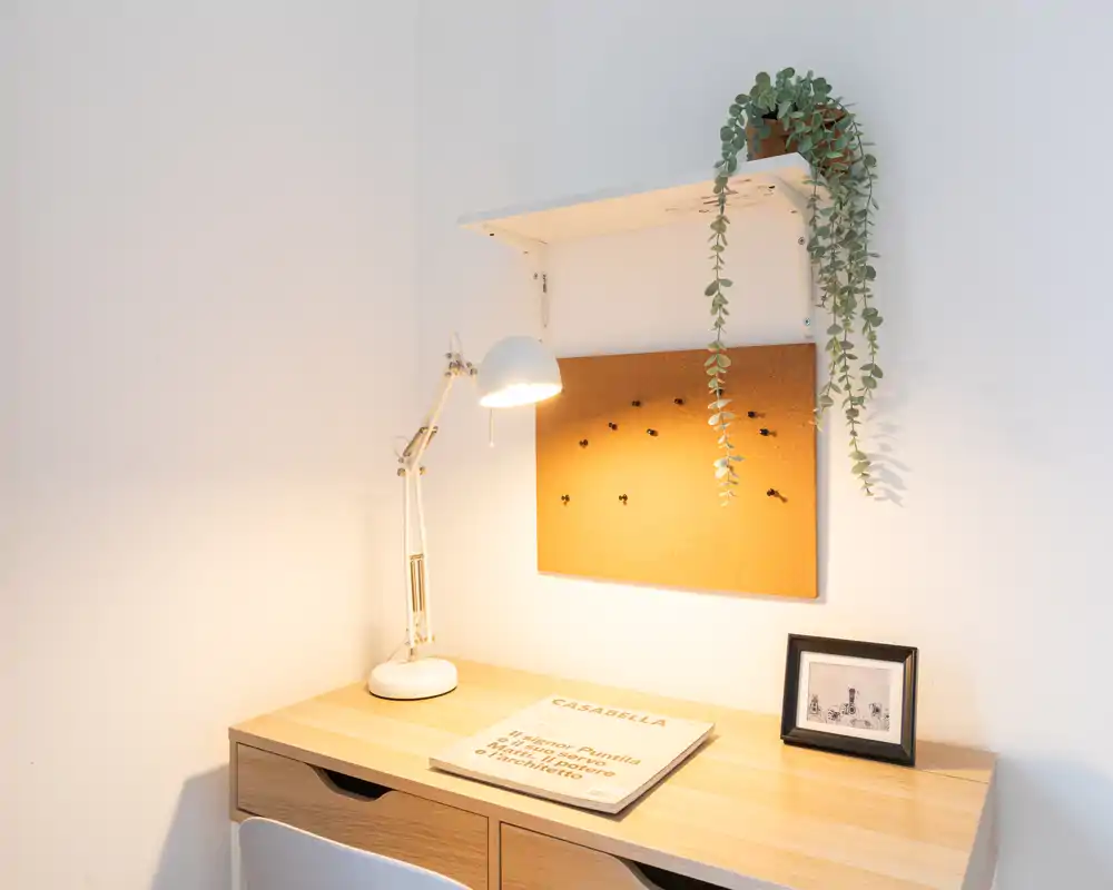 Un rincón de escritorio ordenado con mesa de madera, lámpara de trabajo, corcho y planta colgante — ideal para trabajar o estudiar en un espacio acogedor.