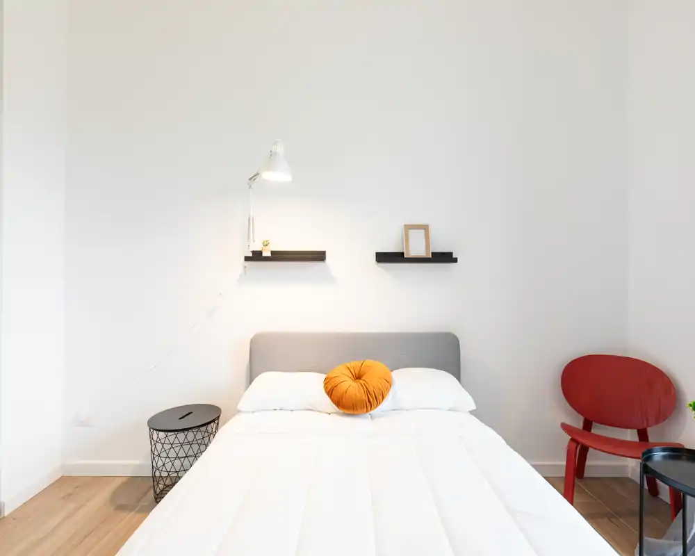 Dormitorio luminoso y minimalista con cama doble centrada, cojín naranja cálido y ropa de cama blanca. Estanterías sencillas y una butaca de diseño aportan un ambiente moderno y relajante.