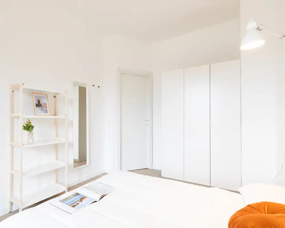 Dormitorio luminoso y minimalista con cama bien hecha, armario blanco y estantería; estilo limpio y aireado perfecto para anuncios modernos.