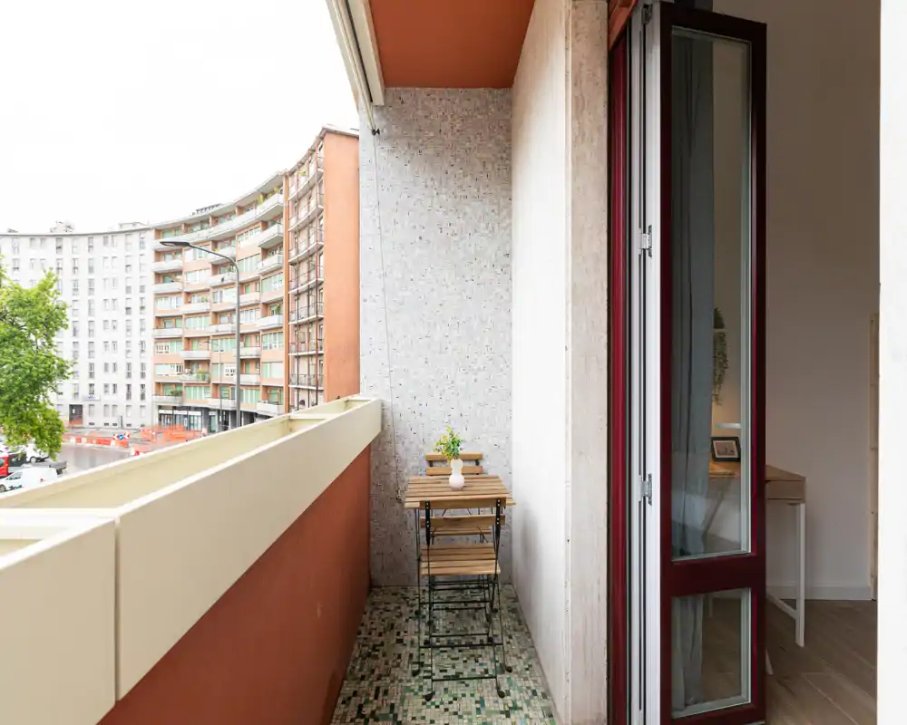 Pequeño balcón amueblado con mesa bistró plegable de madera y dos sillas, suelo en mosaico y vistas a la calle; ideal para el café por la mañana.