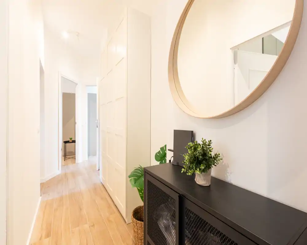 Entrada/pasillo moderno y luminoso con gran espejo circular, consola y planta decorativa; suelo de madera clara y paredes blancas que crean una bienvenida luminosa.