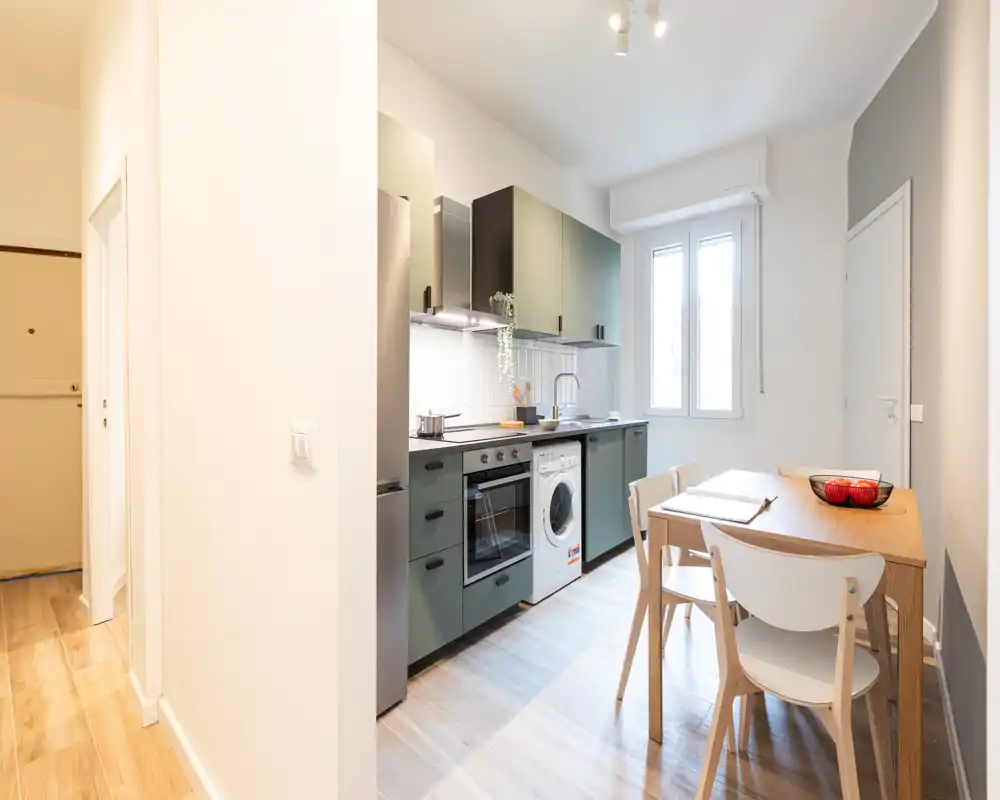 Cocina moderna y compacta con muebles inferiores verde oliva, horno integrado, lavadora y una pequeña mesa de madera junto a una ventana luminosa — práctica y bien cuidada para un apartamento urbano.