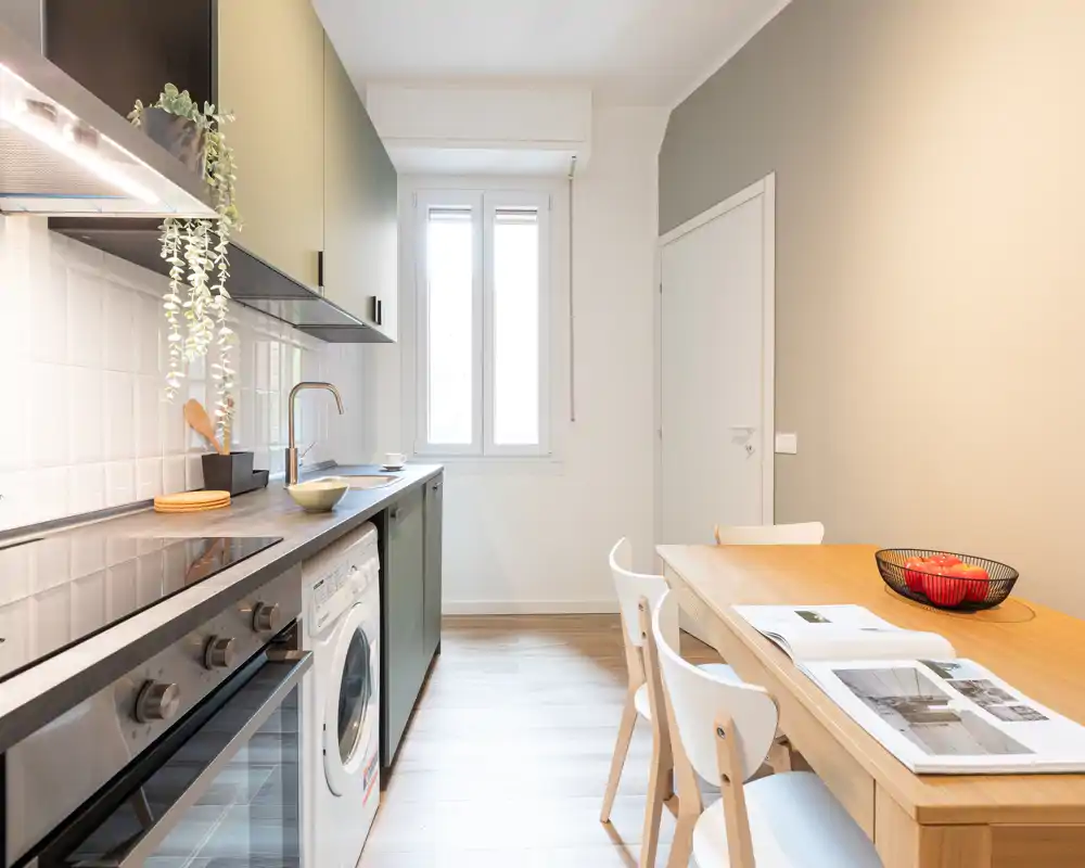 Cocina moderna y luminosa con electrodomésticos integrados, mesa de madera y decoración minimalista. La luz natural de la ventana realza las líneas limpias y la zona de comedor acogedora.