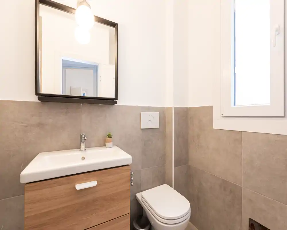 Baño moderno y compacto con inodoro suspendido, mueble de lavabo con espejo, azulejos grises neutros y ventana que aporta luz natural.
