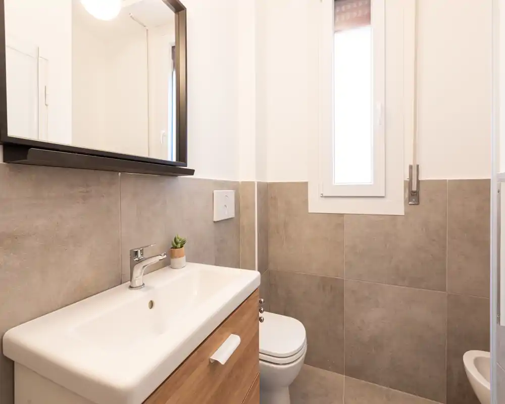Baño moderno y compacto con lavabo de pared y inodoro suspendido, azulejos grises y mueble espejo sobre el lavabo.