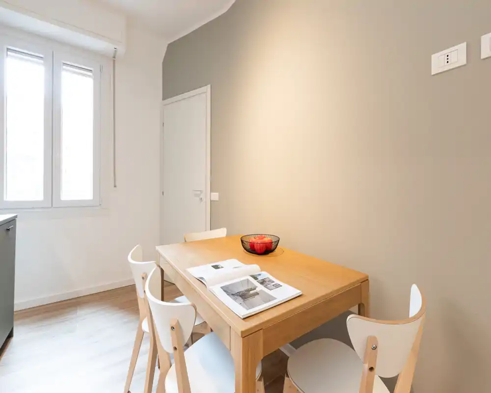 Rincón de comedor luminoso y minimalista con mesa de madera y cuatro sillas blancas, iluminado por una ventana amplia — ideal para comidas informales o un apartamento pequeño.