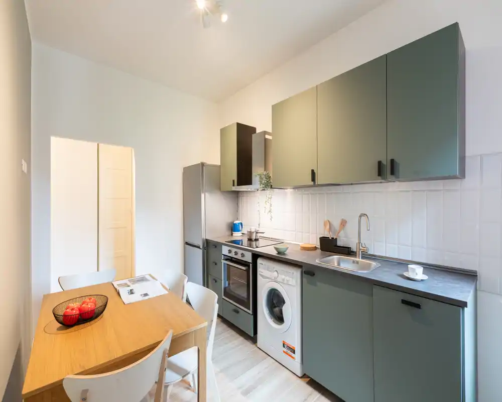 Cocina moderna con armarios verde salvia, horno integrado, lavadora y mesa de comedor de madera para cuatro — luminosa, ordenada y funcional para el día a día.