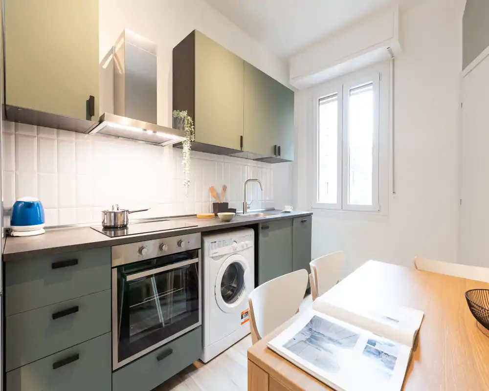 Cocina moderna y luminosa con armarios verde oliva, horno empotrado, lavadora y mesa de madera junto a la ventana — un espacio funcional y ordenado para cocinar y comer de forma informal.