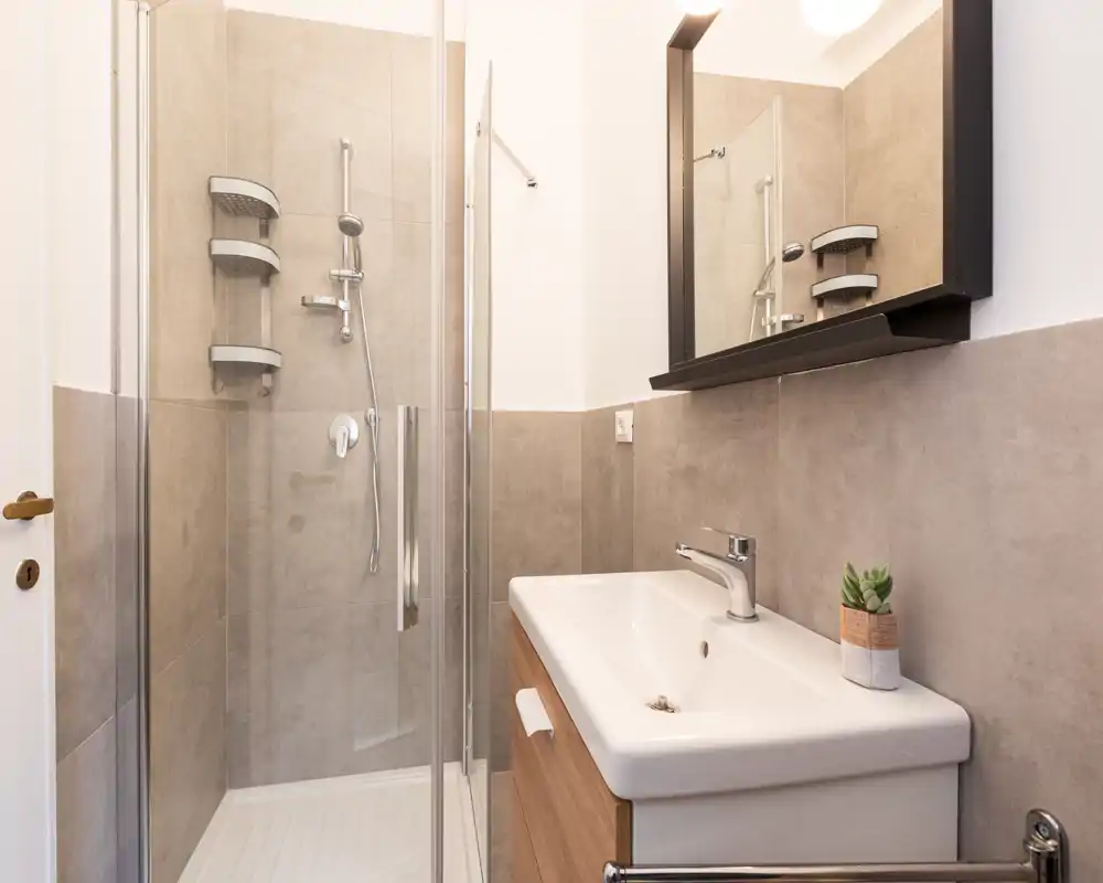 Baño moderno y compacto con ducha de cristal, mueble-lavabo suspendido y espejo con armario. Azulejos neutros y grifería limpia ofrecen un espacio funcional y fresco.