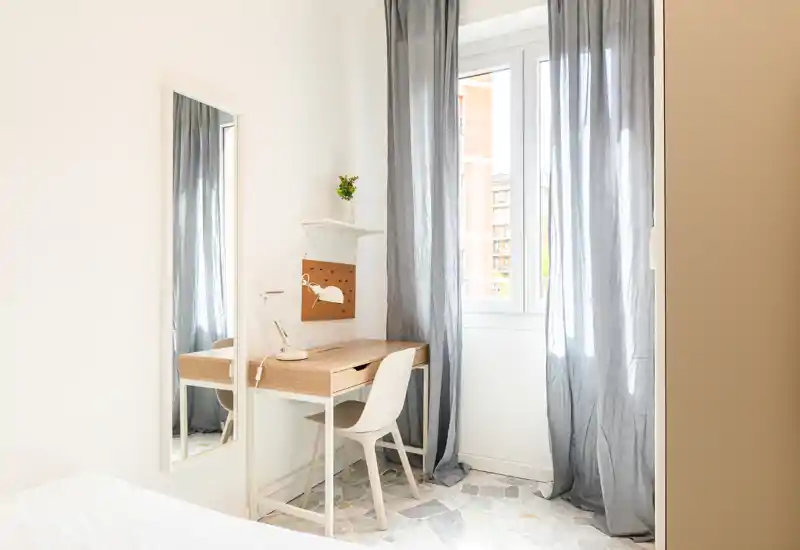 Coin chambre lumineux avec une fenêtre et un espace de travail ; bureau minimal, chaise et miroir pleine longueur offrent une ambiance calme et aérée.