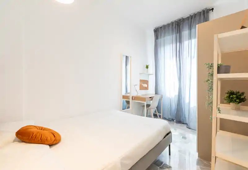 Chambre lumineuse et minimaliste avec lit double, coussin d'accent chaud, petit bureau près de la fenêtre et voilages laissant passer la lumière naturelle.