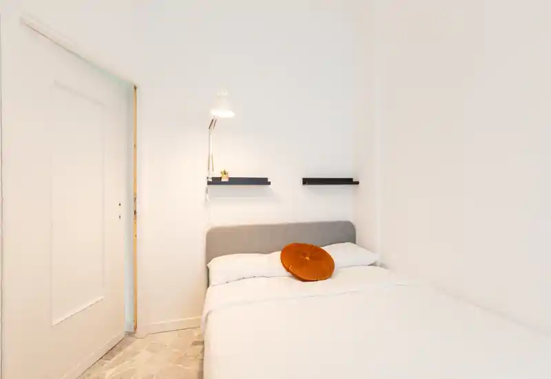 Chambre minimaliste et lumineuse avec un lit double bien fait, tête de lit grise et coussin décoratif orange. Murs blancs, lampe de chevet et étagères flottantes offrent une ambiance moderne et apaisante, parfaite pour la location courte durée.