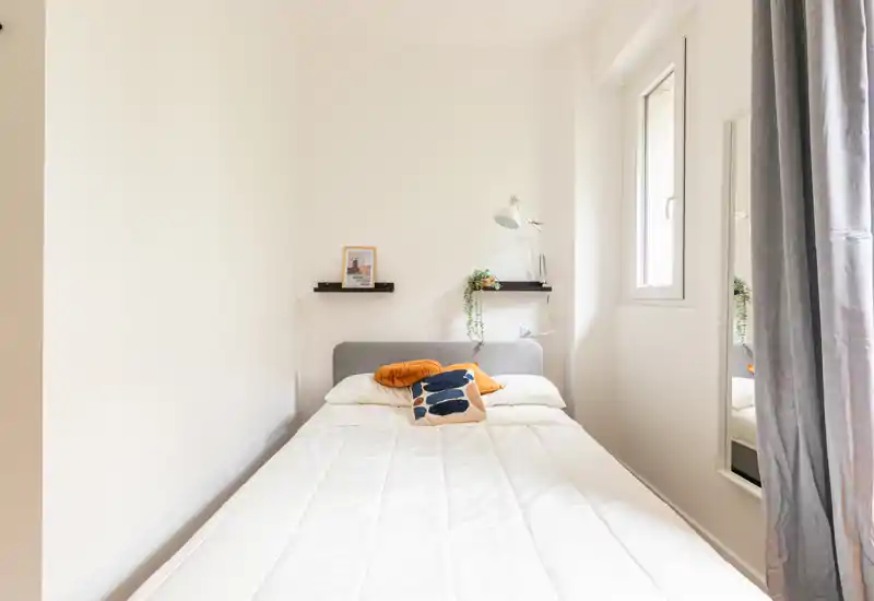 Dormitorio individual luminoso y ordenado con cama cómoda, cojines decorativos, lámpara y plantas; la ventana aporta mucha luz natural.