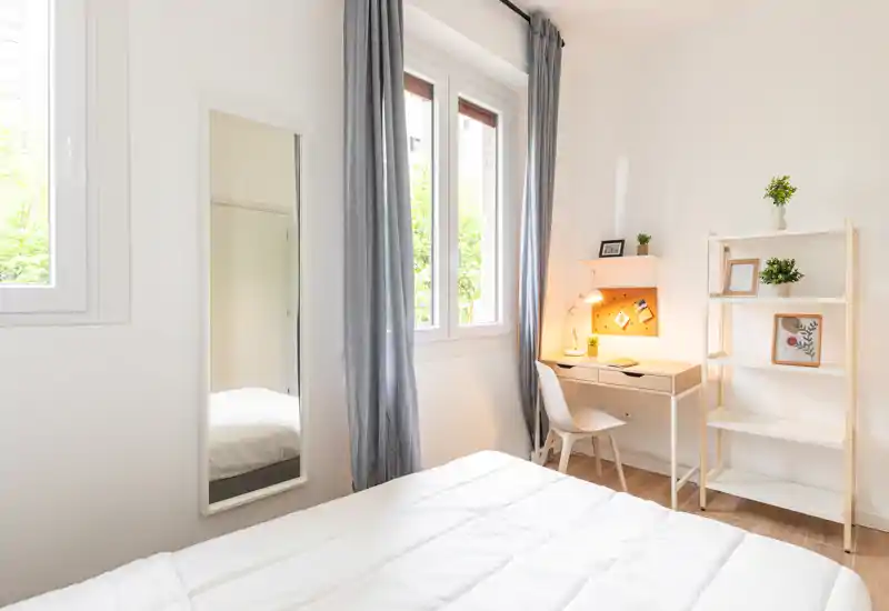 Dormitorio luminoso y minimalista con cama blanca, ventana amplia con cortinas grises, pequeño escritorio con lámpara y estantería — ideal para descansar y trabajar.