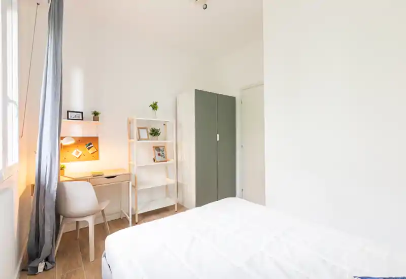 Dormitorio luminoso y minimalista con la cama hecha, un pequeño escritorio y estantería junto a la ventana — ideal para un estudiante o una persona sola.