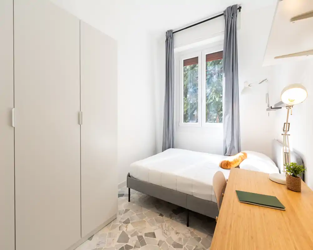 Dormitorio luminoso y minimalista con cama doble, armario y escritorio de madera junto a la ventana: la luz natural y los tonos neutros crean un ambiente tranquilo y aireado.