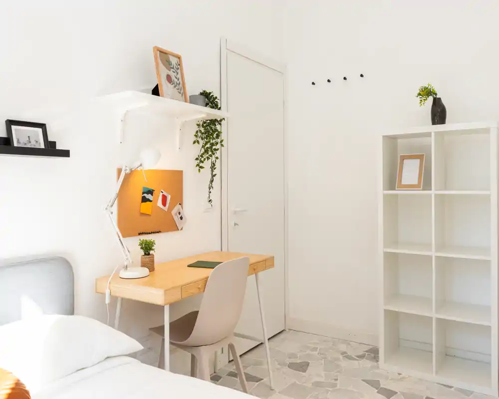 Rincón de dormitorio luminoso y minimalista con borde de cama visible, escritorio de madera con lámpara y silla, estanterías y plantas decorativas — ideal para un alquiler compacto y con estilo.