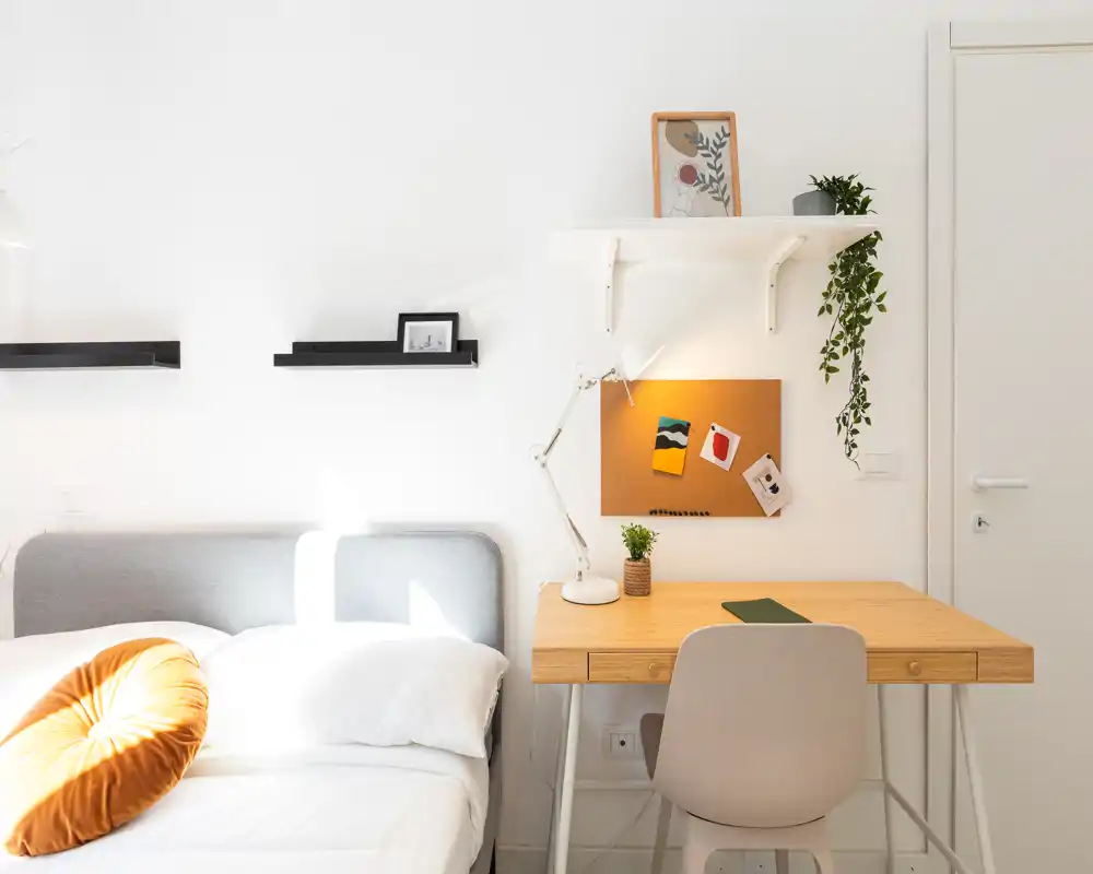 Dormitorio luminoso y minimalista con una cama cómoda, ropa de cama suave y un escritorio de madera ordenado con lámpara — ideal para una estancia moderna y tranquila.