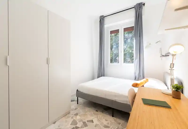 Camera da letto luminosa e moderna con letto matrimoniale, armadio, scrittoio e finestra ampia che lascia entrare luce naturale — linee pulite e tonalità neutre per un ambiente tranquillo.