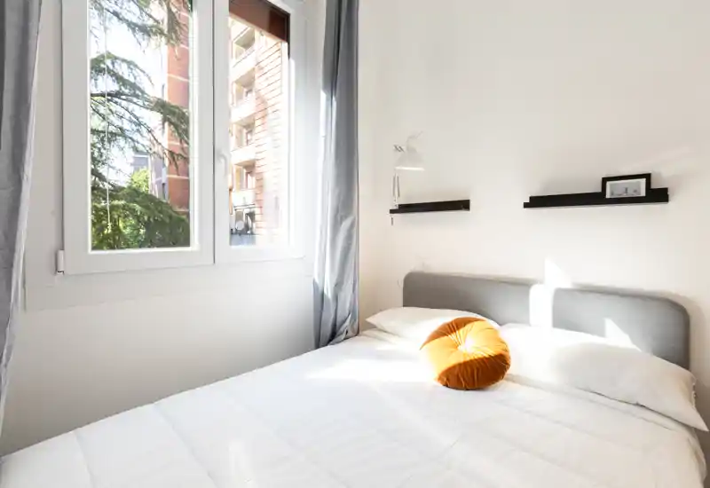 Dormitorio luminoso y minimalista con cama doble cómoda, cabecero gris y un cojín redondo decorativo. La luz natural de la ventana grande crea un ambiente acogedor y aireado.