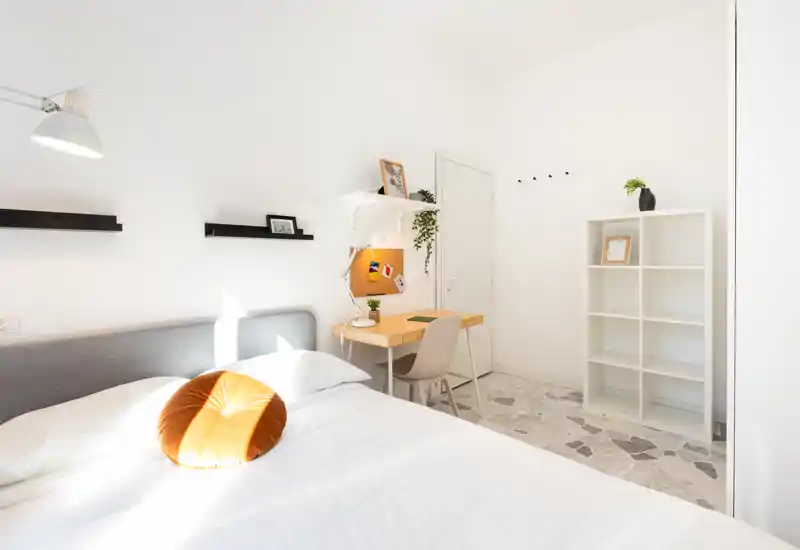 Dormitorio luminoso y minimalista con cama grande, lámpara y pequeño escritorio; paredes blancas y luz natural ofrecen un ambiente tranquilo y aireado.