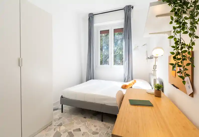 Dormitorio moderno y luminoso con cama doble, escritorio y armario; luz natural desde la ventana y decoración minimalista.
