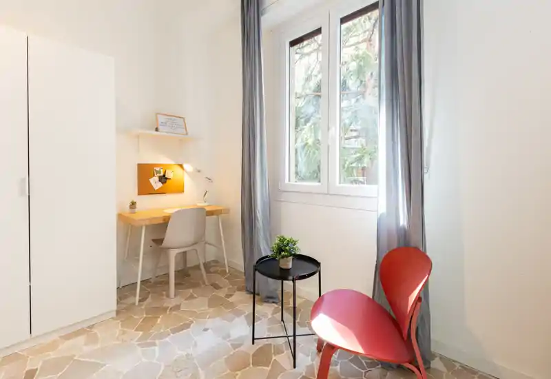 Rincón de dormitorio luminoso y sencillo con escritorio de estudio, armario y sillas junto a la ventana con luz natural.