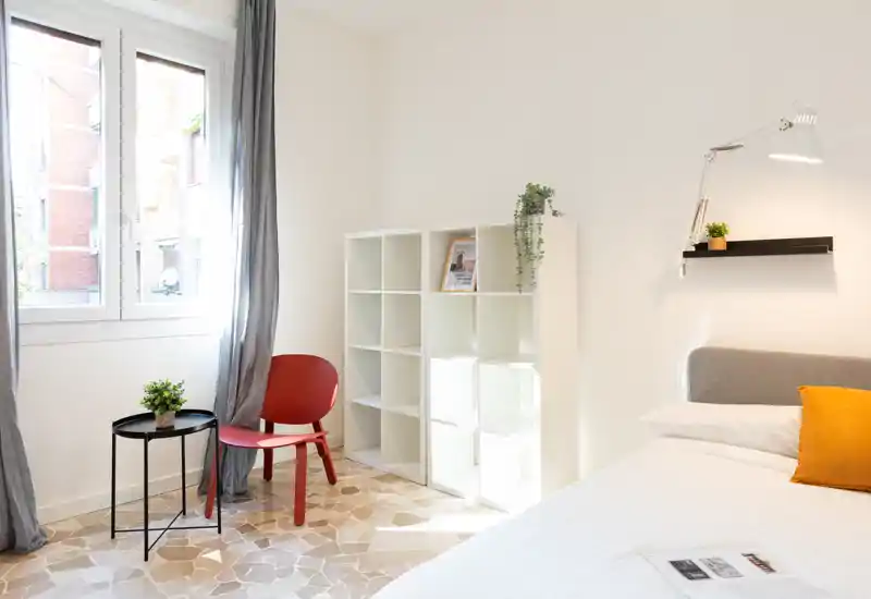 Dormitorio luminoso y minimalista con la cama bien hecha, una estantería moderna y un pequeño rincón con silla junto a la ventana. La luz natural y la decoración sencilla crean un ambiente tranquilo y acogedor.