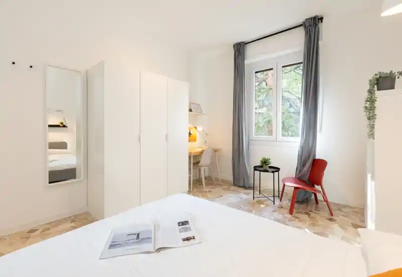 Dormitorio luminoso y minimalista con la cama bien hecha en primer plano, armario blanco, un pequeño escritorio con luz cálida y una ventana que aporta vistas verdes.