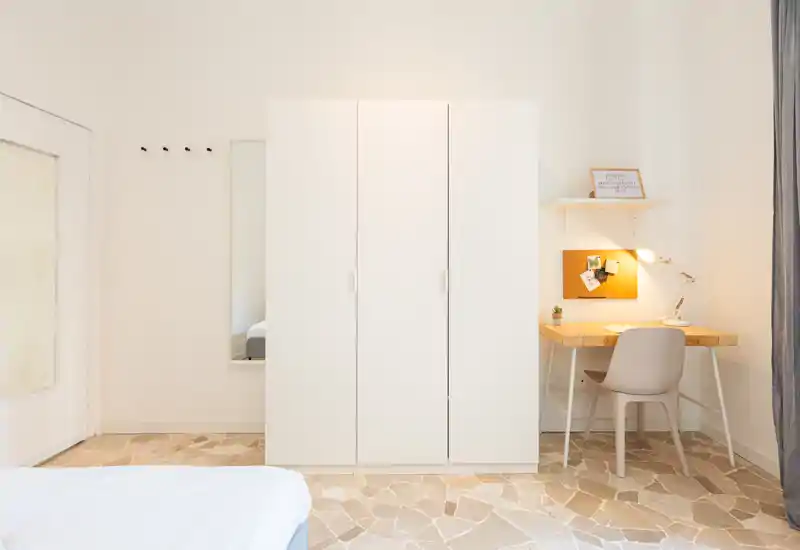 Dormitorio minimalista y luminoso con armario blanco, esquina de la cama visible y un pequeño escritorio de madera con lámpara — espacio limpio y relajante, ideal para estancias cortas.