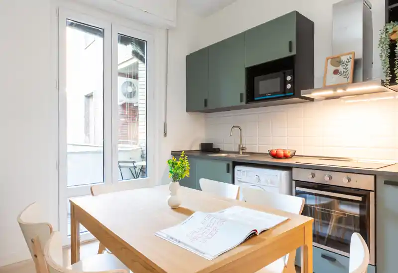 Cocina moderna y luminosa con armarios verdes, horno y microondas integrados, y una mesa de madera junto a una puerta de cristal — perfecta para comidas acogedoras y luz natural.