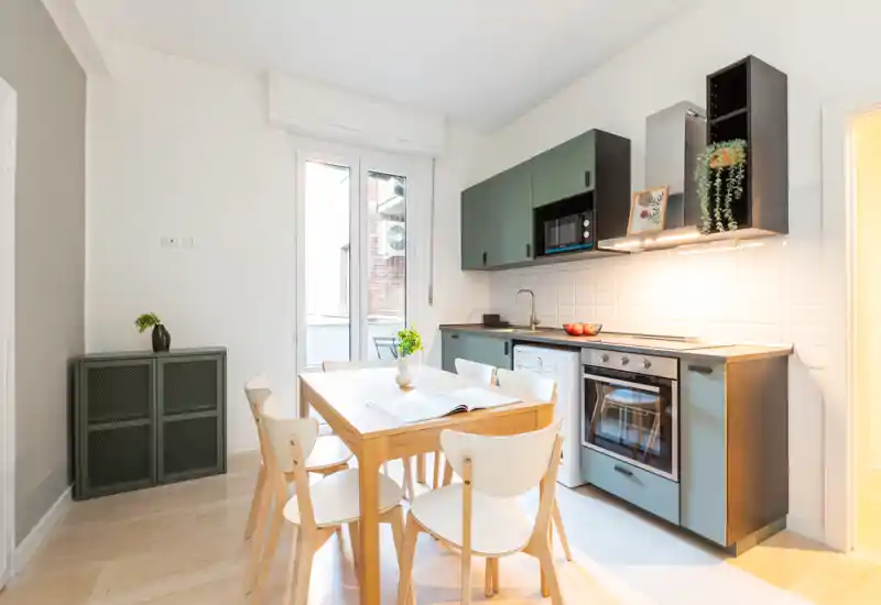 Cocina moderna y luminosa con electrodomésticos integrados y mesa de madera para cuatro; líneas limpias, armarios verdes y luz natural cálida que crean un ambiente acogedor.
