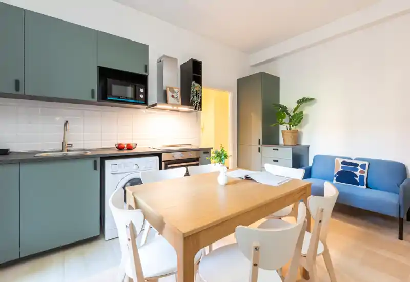 Cocina de planta abierta moderna con armarios verdes, electrodomésticos integrados y mesa de madera para cuatro; luminosa, ordenada y decorada con plantas y cojines para un ambiente acogedor.