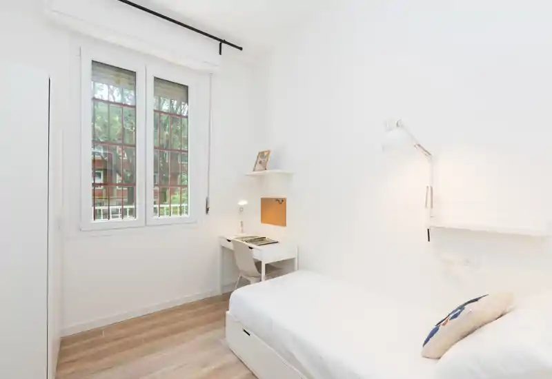 Bright, minimal single bedroom with a neatly made bed, bedside reading lamp and a compact desk by the window — ideal for a student or single occupant.