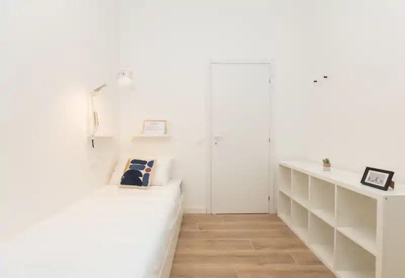 Minimalist single bedroom with a neatly made bed, wall lamp and cubed shelving; bright white walls and warm wood flooring create a calm, modern feel.