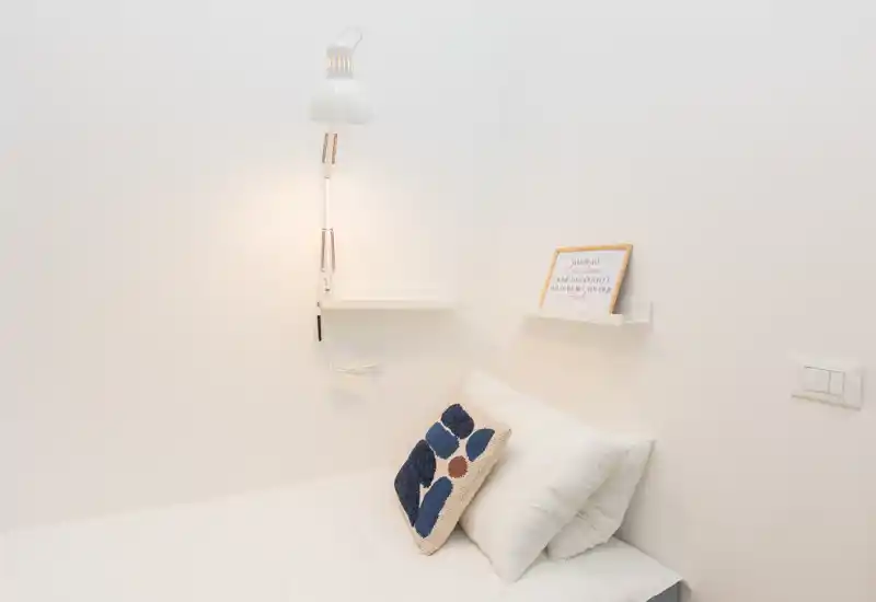 Minimal, bright bedroom corner with a clean white bed, decorative cushion and a wall-mounted lamp creating a calm, stylish atmosphere.