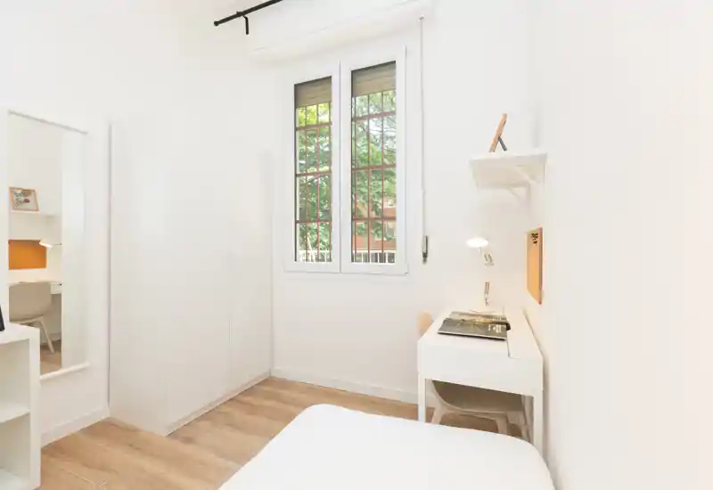 Small bright bedroom with a single bed corner visible, white wardrobe, and a compact desk by the window. Clean minimalist styling and abundant natural light make it ideal for a student or single occupant.